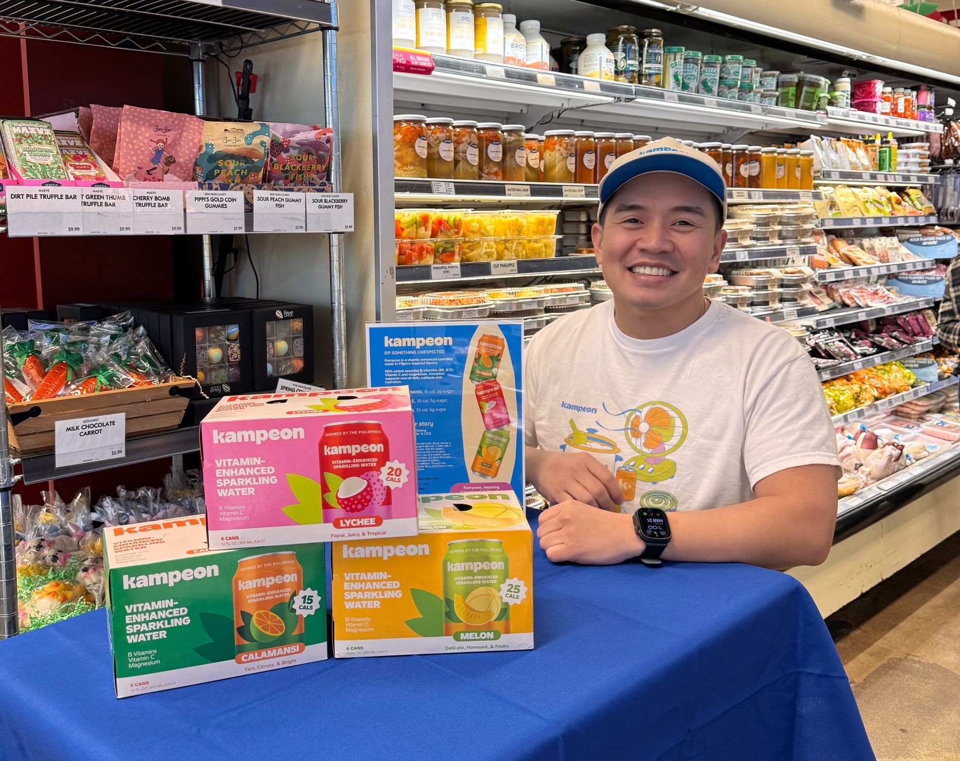 Chris Galang promotes Kampeon vitamin-enhanced sparkling water at a grocery store tasting event, seated behind a table displaying multiple product flavors and brand information.