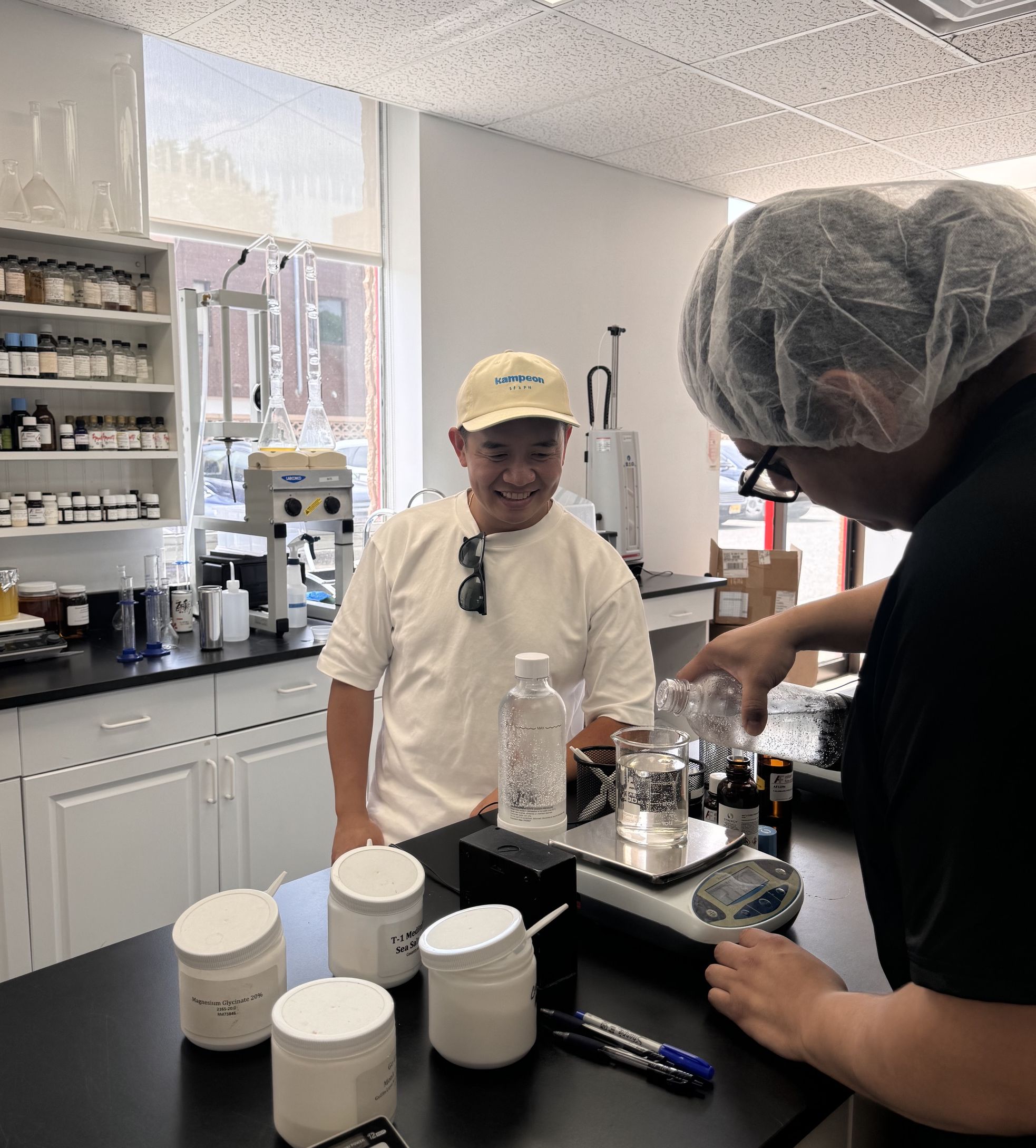 Chris Galang visits a beverage development laboratory, reviewing product formulation and testing alongside a team member at a workbench equipped with scales, glassware, and ingredient samples.
