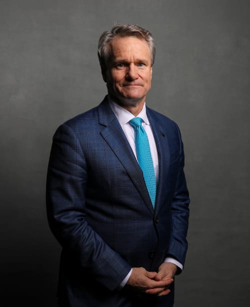 Brian Moynihan wearing a dark blue suit and a light blue tie, with his hands united in front of his body.